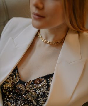 A stylish woman wearing a gold necklace and embellished top with a white blazer indoors.